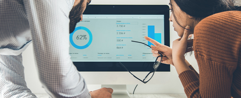 Two people are looking at analytics graphics on a computer, a man who is typing and a woman who is pointing to the screen with her glasses