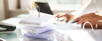 A stack of receipts impaled on a paper nail in the foreground, a hand holding a pen in the background