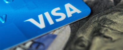 The Visa logo on a blue credit card sitting atop a $100 bill